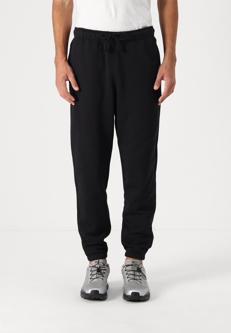 Zwarte trainingsbroek van zacht stof, met een elastische tailleband en trekkoord, taps toelopende enkels en zijzakken. Draagt met grijze sportschoenen.