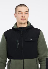 Schwarze und grüne, fleecegefütterte Jacke mit Kapuze, Reißverschluss vorne, zwei Seitentaschen und einer Brusttasche; mit Logo-Details und kontrastierenden Texturen.