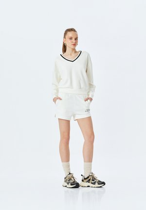 Weißer Kapuzenpullover mit schwarzem V-Ausschnitt, kombiniert mit weißen Shorts mit seitlichen Schlitzen. Tragen mit schwarz-beigen Sneakern und hellen Socken.