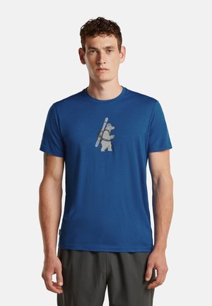 Mann trägt ein blaues T-Shirt mit einer Grafik eines Bären, der Skier und einen Wanderstock vorne trägt, steht vor einfachem Hintergrund.
