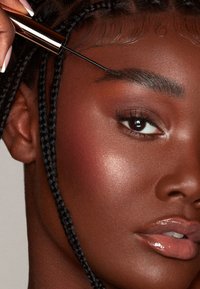 Applicateur de gel à sourcils maintenu près de cheveux tressés foncés ; la peau affiche un fini lisse et éclatant avec des reflets subtils ; couleur de lèvres brillante et neutre.