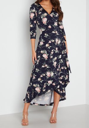 Woman wearing a navy blue floral wrap dress with asymmetrical hem and three-quarter sleeves, paired with clear strap heels.