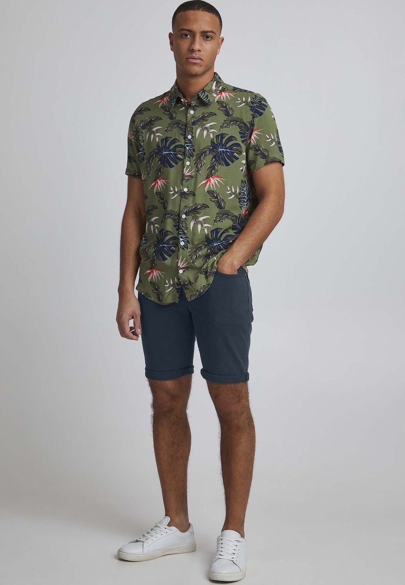 Kortermet grønn skjorte med tropisk bladmønster, parret med oppbrettede marineblå shorts og hvite joggesko. Enkel design med knapper foran.