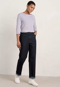 Pruhovaný dlouhý rukáv v levandulové a bílé barvě, v kombinaci s džínami vysokého pasu v tmavé barvě a bílými teniskami. Modelka stojí, ruce má v kapsách.