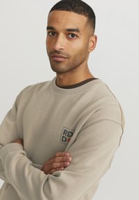 Light beige sweatshirt with a crew neck, featuring a small graphics patch in the chest area. Soft fabric and relaxed fit.