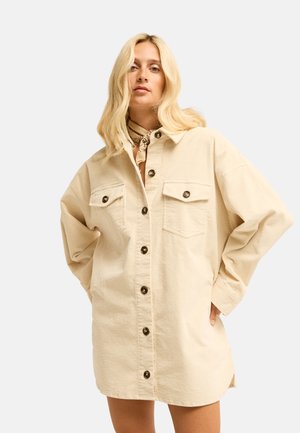 Vrouw met blond haar die een beige corduroy blousejurk met knopen draagt en een patroonige halssjaal tegen een effen achtergrond.