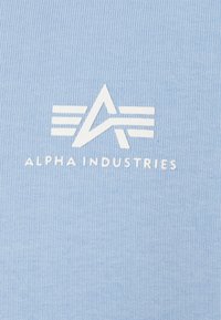Ανοιχτό γαλάζιο βαμβακερό t-shirt με λευκό λογότυπο που περιέχει ένα στυλιζαρισμένο γράμμα "A" και τρεις γραμμές από κάτω, με το κείμενο "ALPHA INDUSTRIES" κάτω.