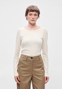 Μακρυμάνικο τοπ σε κρέμα από μαλακό ύφασμα με στρογγυλή λαιμόκοψη, σε συνδυασμό με ταμπά παντελόνι που διαθέτει κουμπί και μπροστινές τσέπες.