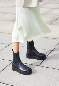 Black leather ankle boots with a rounded toe and elastic side panels, featuring a thick platform sole. Worn with a flowing light-colored skirt.