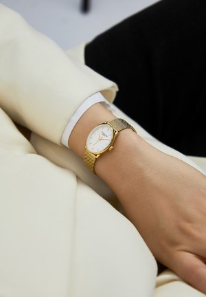 Sat zlatne boje s okruglim bijelim brojčanikom, mrežastim remenom i minimalističkim dizajnom. Nosi se na zapešću, uparen s kremastim odjevnim predmetom.