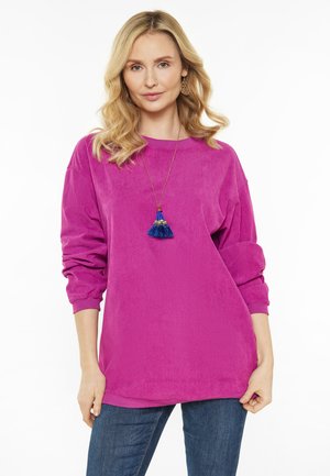 Lys pink corduroy top med lange ærmer, rund halsudskæring og ribbet tekstur. Båret med en blå kvast halskæde og denim jeans.