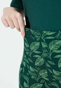 Leggings verts avec un motif de feuilles en vert clair. Tissu lisse, design ajusté, taille élastique, montrés en train d'être pincés à la taille.