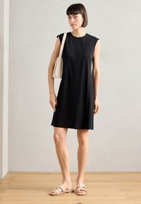 Black sleeveless dress with a round neckline, made of smooth fabric. Worn with a beige shoulder bag and light pink sandals.