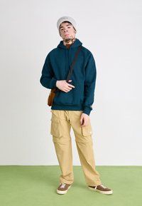 Dunkelblauer Hoodie mit Fronttasche, beige Cargo-Hosen mit seitlichen Taschen und braunen Sneakers mit weißen Sohlen, ergänzt durch eine helle Mütze.