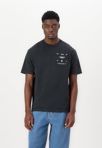 Black cotton t-shirt with short sleeves, round neckline, featuring white graphic text and design on the front, paired with blue jeans.