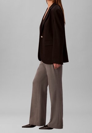 Frau trägt schwarzen Blazer, graue weit geschnittene Hose und spitze dunkelbraune Schuhe, steht seitlich vor einem einfarbigen grauen Hintergrund.