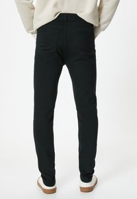 Jeans elásticos negros con un ajuste slim, diseño de cinco bolsillos y detalles de costura sutiles, combinados con zapatillas blancas y una blusa beige.