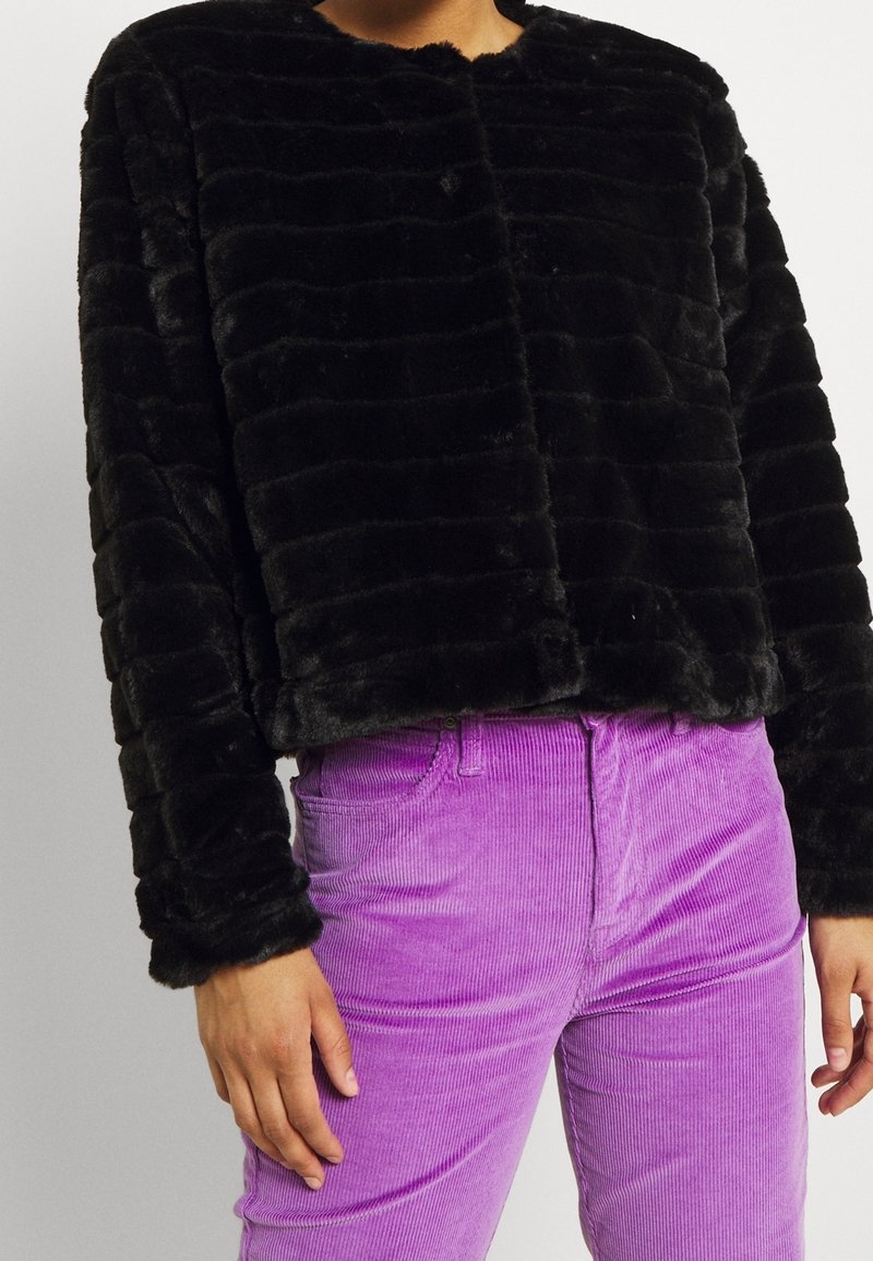 Chaqueta corta de piel sintética negra con rayas horizontales, con escote redondo y mangas largas, combinada con pantalones de pana color lila claro.