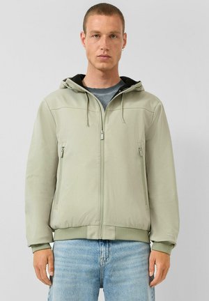 Mann trägt eine hellgrüne Kapuzenjacke mit Reißverschluss und zwei Reißverschlusstaschen, steht vor einem einfarbigen grauen Hintergrund.