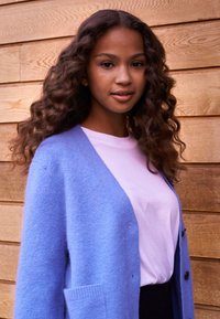 Light blue cardigan made of soft fabric, paired with a pale pink shirt. Wooden background with horizontal planks adds contrast.