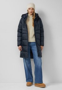 Manteau matelassé bleu marine avec capuche, affichant des coutures horizontales. Porté par-dessus un pull crème et un jean large bleu ; complété par des bottes tan.