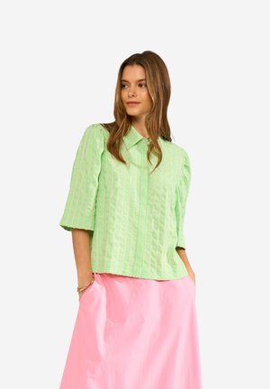 Kobieta ubrana w jasnozieloną, paskowaną bluzkę z bufkami i różową spódnicę, stojąca z rękami w kieszeniach na białym tle.