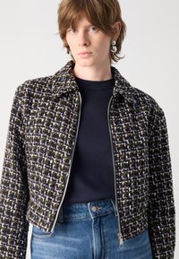 Giacca tessuta in tonalità scure con un motivo testurizzato, caratterizzata da una chiusura a zip, un colletto a punta e accessori in tonalità argentata. Abbinata a un top in maglia blu.