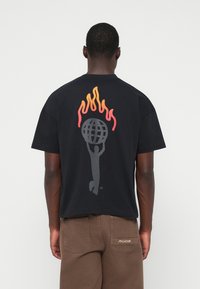 Άνδρας που φορά μαύρη μπλούζα με γραφικό σχέδιο ενός ατόμου που κρατάει μια σφαίρα περικυκλωμένη από πορτοκαλί-κόκκινες φλόγες στην πλάτη, συνδυασμένη με καφέ παντελόνι.