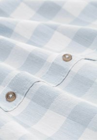 Tissu bleu clair à carreaux avec des carrés alternés. Présente de petits boutons ronds de couleur marron clair et une surface douce et texturée.