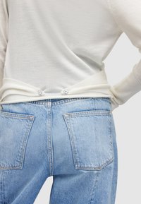 Weißes Langarmoberteil mit gerippten Bündchen, kombiniert mit hellblauen hochgeschnittenen Jeans. Das Oberteil hat zwei Knöpfe hinten.