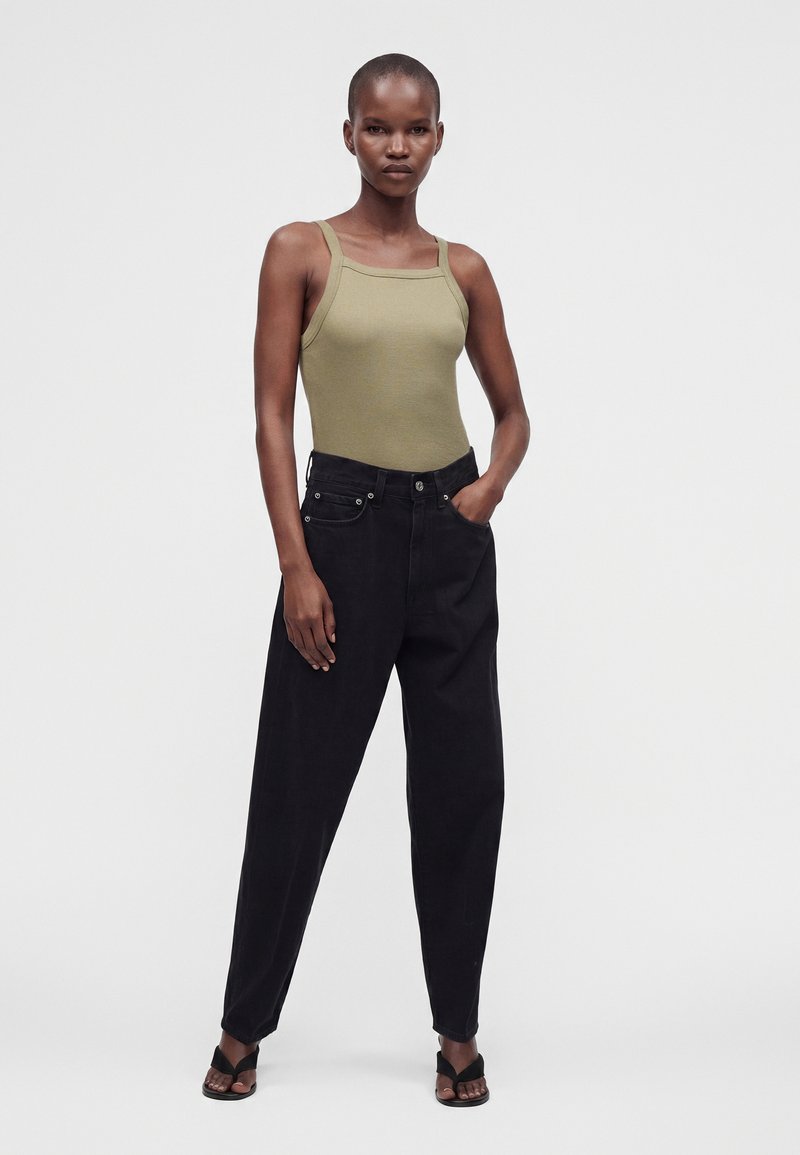 Grande femme portant un haut sans manches vert olive, un pantalon noir ample taille haute et des sandales noires à bout ouvert, debout avec une main dans la poche.