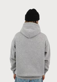 Felpa grigia con cappuccio realizzata in tessuto morbido, caratterizzata da una vestibilità comoda, tasca a marsupio e polsini a costine. Indossata con un berretto nero.