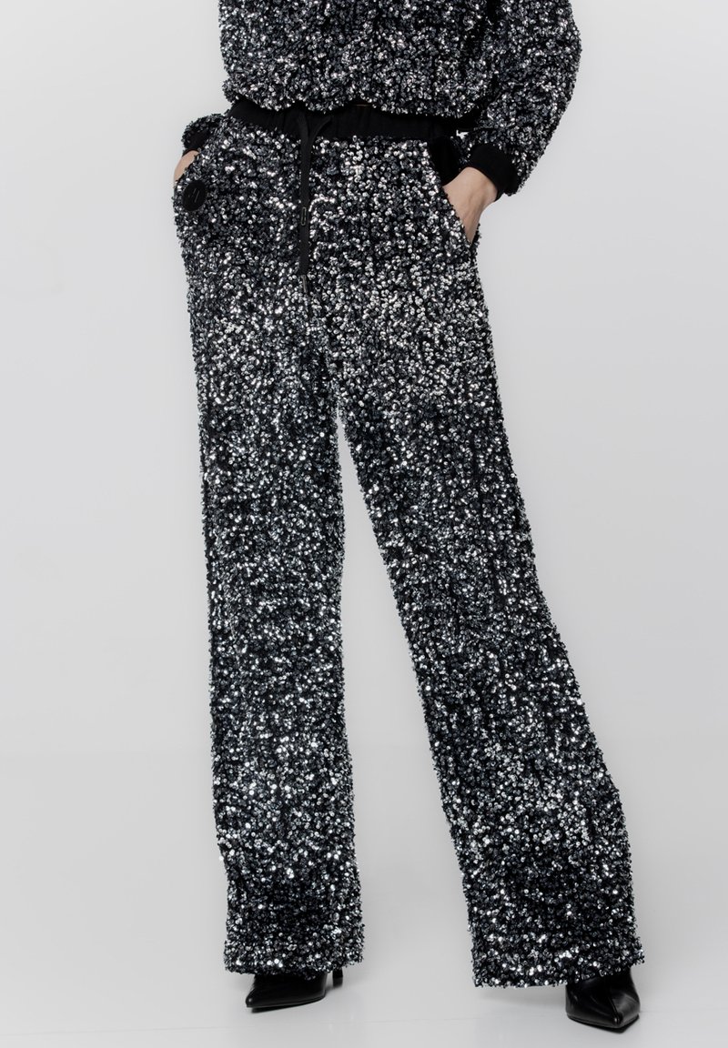 Czarne i srebrne spodnie z szerokimi nogawkami ozdobione cekinami, z teksturowaną powierzchnią, posiadające elastyczny pas i boczne kieszenie, stylizowane na z wysmuklającymi czarnymi butami.