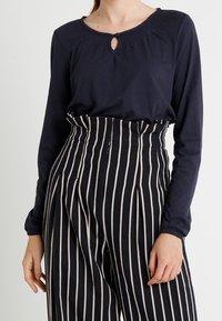 Tmavě modrý top s dlouhým rukávem a detailem výřezu u krku, spojený s vysokým pasem a vertikálně pruhovanými kalhotami v černé a krémové barvě.