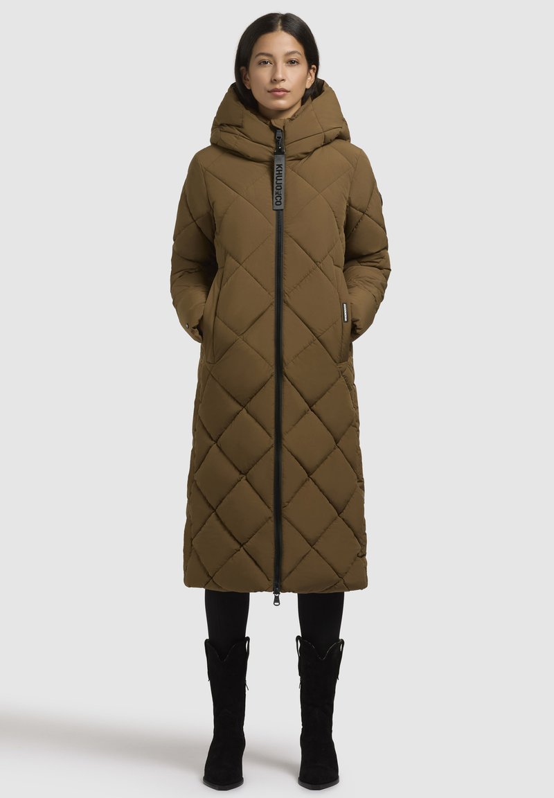 Cappotto lungo marrone trapuntato con cappuccio, dotato di zip frontale, tasche laterali e motivo a rombi textured. Stivali neri completano il look.