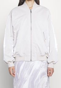 Lehká bílá bomber bunda na zip vpředu, s žebrovanými manžetami a lemem. Má boční kapsy a hladkou, lesklou texturu.