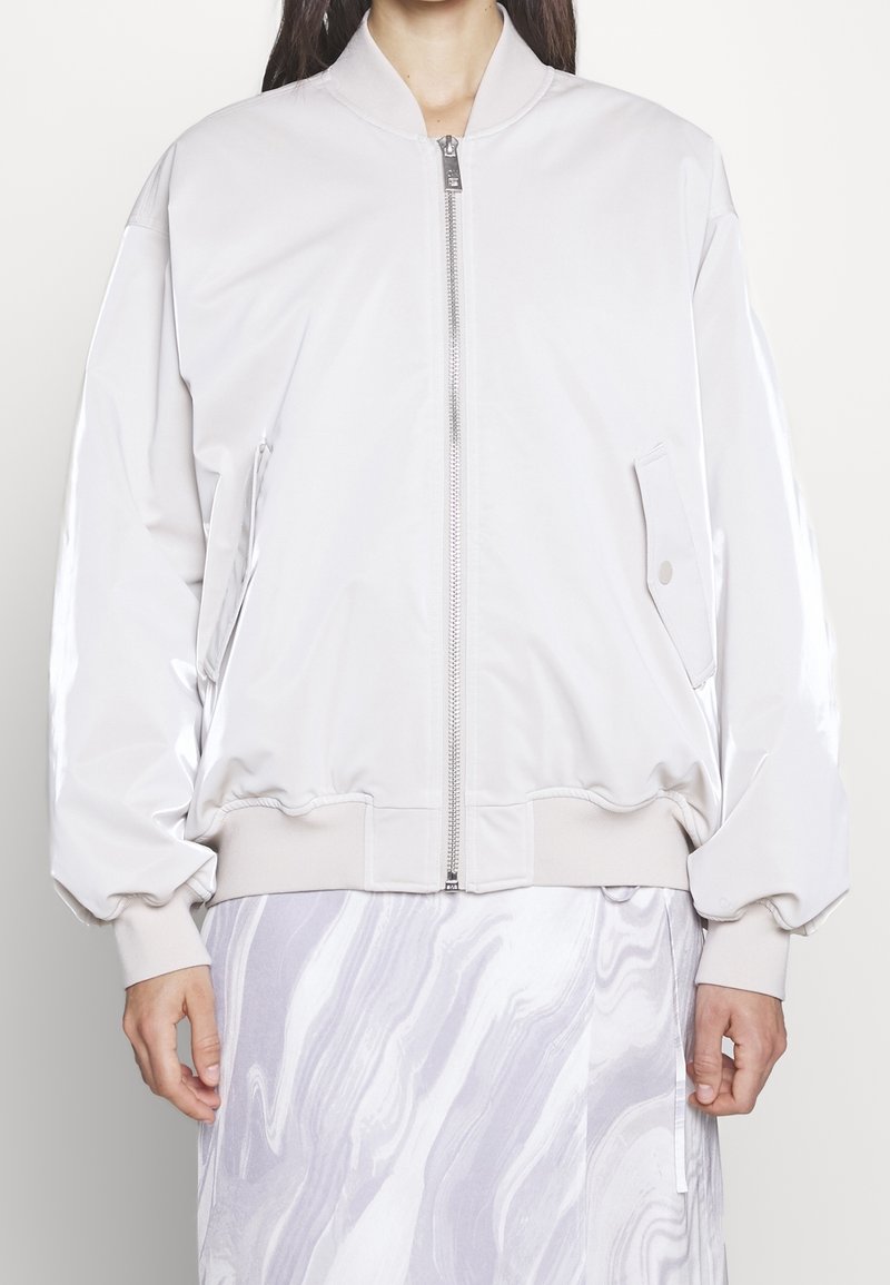 Lehká bílá bomber bunda na zip vpředu, s žebrovanými manžetami a lemem. Má boční kapsy a hladkou, lesklou texturu.