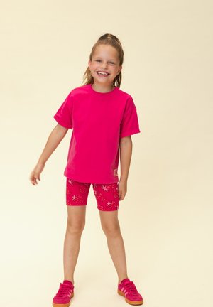 Lachend meisje dat staat, draagt een felroze oversized T-shirt, rode korte broek met aardbeienprint en bijpassende roze sneakers op een beige achtergrond.