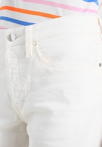 Jeans de mezclilla blanca con un diseño clásico de cinco bolsillos y un cierre de botón plateado. La textura se muestra suave con un ajuste estructurado.