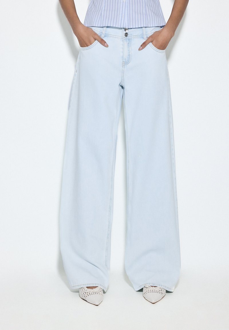 Personne portant un jean large bleu clair, mains dans les poches, associé à des chaussures blanches tissées à bouts pointus et une chemise rayée rentrée.
