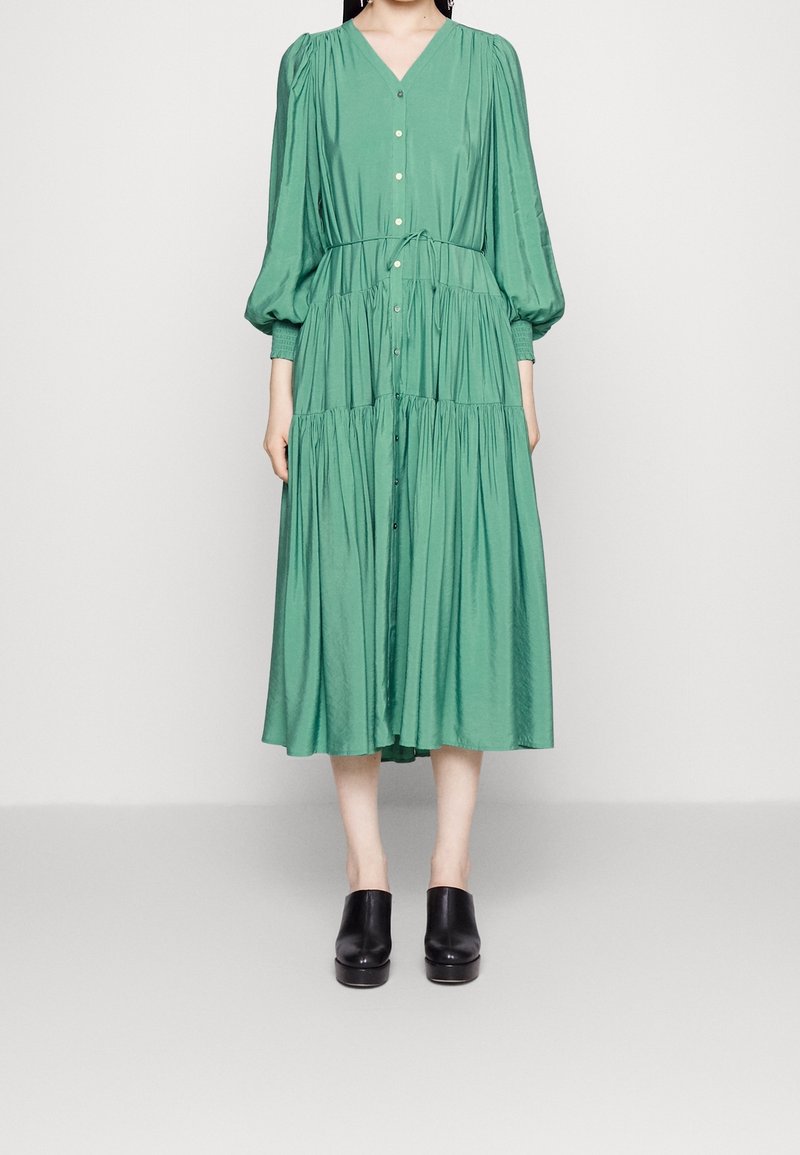 Femme portant une robe midi verte ample à manches longues, boutonnée, ceinturée à la taille, et des chaussures noires sans lacets, debout devant un fond uni.