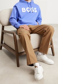 Ljusblå hoodie med vit "BOSS"-logotyp, parat med beige byxor och vita sneakers. Sitter på en trästol med en stoppad beige sits.