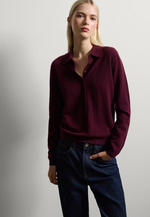 Pullover in magenta con collo a camicia, apertura con bottoni e polsini a coste, abbinato a jeans in denim blu scuro. Tessuto liscio, leggermente elasticizzato.