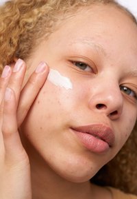 Crème blanche appliquée sur la joue, peau claire avec des imperfections, couleur naturelle des lèvres, cheveux blonds ondulés, fond neutre.