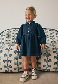 Denimkjole med lange ballonærmer, knaplukning foran og en lomme. Båret med mønstrede sneakers og røde sokker, stillet op imod en blomstret sofa.