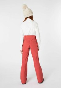 Rote Schneehosen mit hohem Bund und ausgestellten Beinen, kombiniert mit einem weißen Langarmoberteil und einer cremefarbenen Strickmütze.