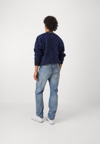 Marineblauer Pullover mit Rautenmuster, kombiniert mit hellblauen Jeans und weißen Sneakern. Der Pullover hat gerippte Bündchen und einen gerippten Saum.