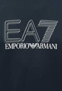 Tmavá látka s bílým obrysem textu "EA7" nad nápisem "EMPORIO ARMANI" a logem orla značky umístěným mezi těmito slovy.