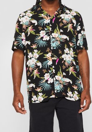 Mann trägt ein schwarzes Kurzarmhemd mit Knöpfen und weißem, pinkem und grünem Blumenmuster sowie schwarze Shorts.