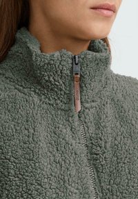 Fleecejakke i dæmpet grøn med høj krave, struktureret overflade, lynlåslukning og trækstrop i brunt læder. Komfortabelt design for varme.